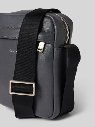 Produktfoto, Handtasche mit Label-Detail Modell 'RAISED REPORTER' von CK Calvin Klein in Dunkelgrau mit Hauptfach, Reißverschluss und Schulterriemen, Breite 17 cm, Länge 20 cm, Tiefe 7 cm, Riemenlänge 68 cm, Herren Accessoire, feine Narbung, silberfarbene Details.