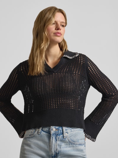 Modelka kobieca nosi Slim Fit Poloshirt in Strick-Optik Modell 'CAITLYN' od Noisy May, bawełniana mieszanka, długi rękaw, półprzezroczysty design, kolor czarny, widoczne drobne dzianinowe oczka, kołnierzyk polo, krój slim fit, widok całej sylwetki z przodu.