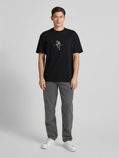 Männermodel trägt Regular Fit T-Shirt aus reiner Baumwolle Modell 'DOLLO' von HUGO, unifarben schwarz mit grafischem Muster, Ganzkörperansicht von vorne.