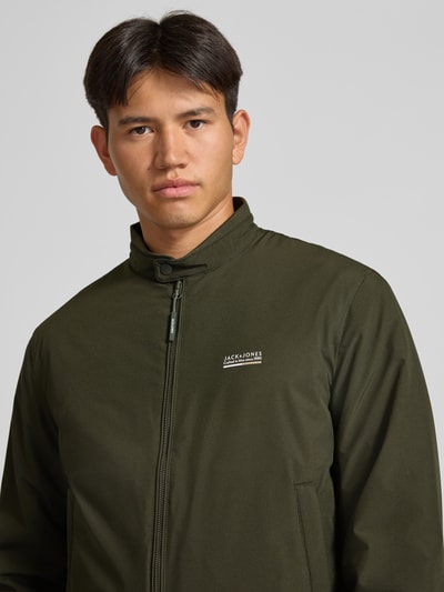 Männermodel trägt Bomberjacke mit Logo Modell 'BROOKLYN' von JACK & JONES in Olivgrün, Reißverschluss, Nahaufnahme Oberkörper. Regular Fit, glatte Textur, Stehkragen.