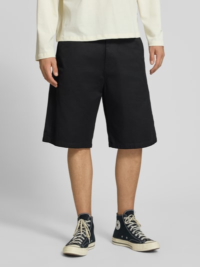 Männermodel trägt Bermudas mit Baumwoll-Anteil Modell 'Brader' von Carhartt Work In Progress, schwarzer Stoff, Relaxed Fit, Nahaufnahme Unterkörper, Produktdetails: Material-Mix, Knopf- und Reißverschluss, Eingriff- und Gesäßtaschen, Gürtelschlaufen, Bundweite 44 cm, Innenbeinlänge 30 cm. Ganzkörperansicht von vorne.