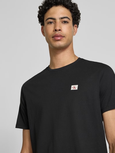 Männermodel trägt T-Shirt mit Label-Badge von Calvin Klein Jeans in Schwarz, reine Baumwolle, Regular Fit, Nahaufnahme Oberkörper.