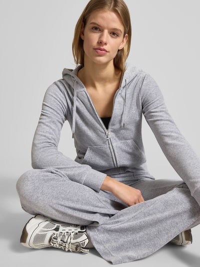 Frauenmodel trägt graue Sweatjacke mit Label-Stitching Modell 'ROBERTSON' von Juicy Couture aus Material-Mix mit Stretch-Anteil, Regular-Fit, Kapuze mit Tunnelzug, Reißverschluss und Känguru-Tasche, Ganzkörperansicht von vorne.