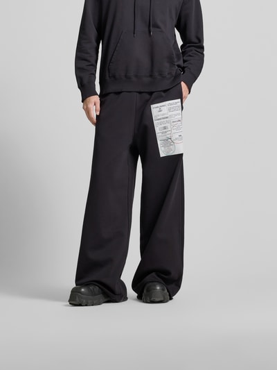 Männermodel trägt Loose Fit Stoffhose mit Logo-Print von MM6 Maison Margiela in Schwarz, reine Baumwolle, elastischer Bund, seitliche Taschen, Nahaufnahme Unterkörper.