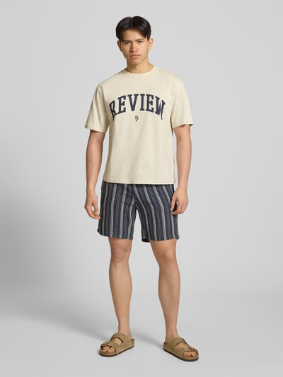 Męski model nosi Shorts mit elastischem Bund und Logo-Detail von REVIEW, bawełniany materiał, krój regular fit, szerokość w rozmiarze M 42 cm, długość nogawki wewnętrznej 20 cm, paski w kolorach granatu, beżu i szarości, widok całej sylwetki z przodu.