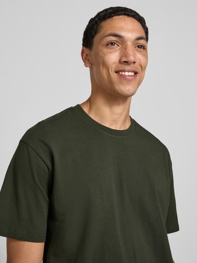 Męski model nosi Regular Fit T-Shirt mit Rundhalsausschnitt od Jack & Jones, czysta bawełna, krój regular fit, okrągły dekolt, rękaw o dł. 1/2, długość tyłu w rozmiarze M: 70 cm, kolor ciemnozielony, gładka faktura. Zbliżenie na górną część ciała.
