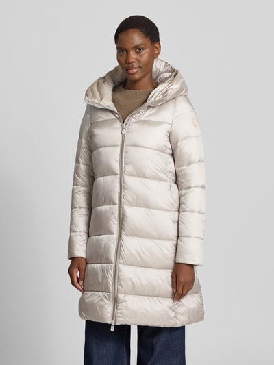Frauenmodel trägt Steppmantel mit Kapuze Modell 'LYSA' von Save The Duck in Beige mit Zweiwege-Reißverschluss, Ganzkörperansicht von vorne.