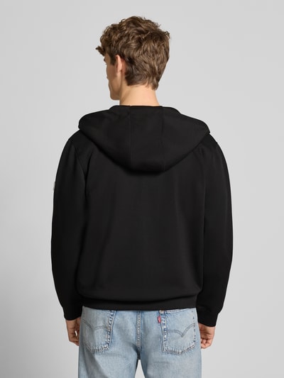 Männermodel trägt Sweatjacke mit Reißverschluss von Guess in Schwarz, Baumwoll-Materiaalmix, los model, capuchon, steekzakken, ribboorden, ruglengte 66 cm, mouwlengte 66 cm, ganzkörperansicht rückansicht.