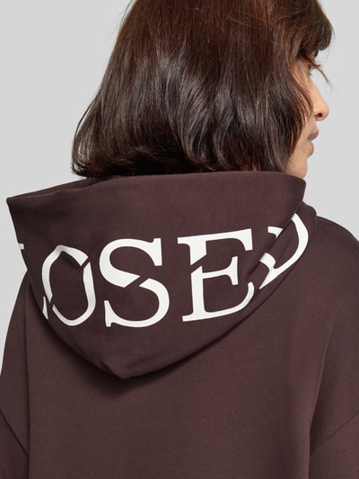 Frauenmodel trägt Oversized Hoodie aus Baumwolle von Closed in Dunkelbraun mit weißem Print und überschnittenen Schultern, Nahaufnahme Rückansicht.