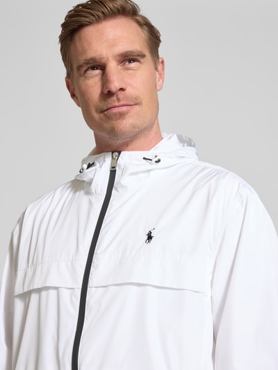 Mężczyzna nosi Regular Fit Regenjacke mit Logo-Stitching von Polo Ralph Lauren, efekt: jednolity kolor, kaptur z tunelem, zamek błyskawiczny dwustronny, długi rękaw, elastyczne zakończenie rękawów, wyhaftowane logo, wpuszczane kieszenie, krój regular fit, zbliżenie na górną część ciała