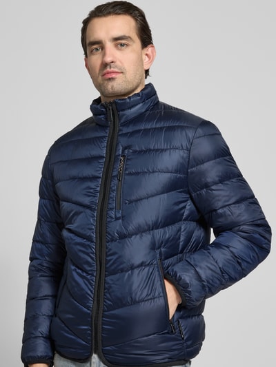 Mężczyznamodel nosi Regular Fit Steppjacke mit Stehkragen von Tom Tailor w kolorze granatowym, krój regular fit, stójka, długi rękaw, elastyczne wykończenia, wodoodporna, kieszenie na zamek, naszywki, widok na całe ciało z przodu.