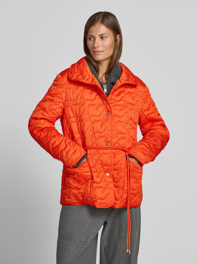 Vrouwenmodel draagt feloranje gewatteerd jack met opstaande kraag, knoopsluiting, opgestikte zakken, strikceintuur van Marc Cain, regular fit, ruglengte maat M 70 cm, schouderbreedte 45 cm, borstwijdte 43 cm, mouwlengte 62 cm, geheel van voren.
