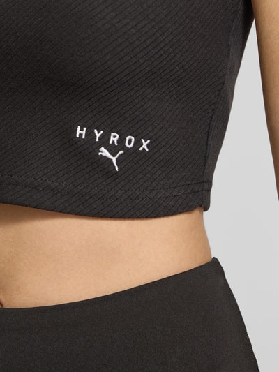 Frauenmodel trägt PUMA x HYROX - Crop Top mit Label-Stitching aus schwarzem Baumwoll-Mix mit Stretch, feine Rippstruktur, Nahaufnahme Oberkörper.