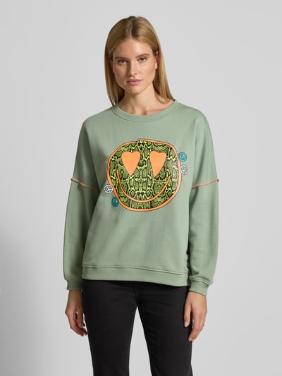 Frauenmodel trägt Oversized Sweatshirt mit Motiv-Print Modell 'Heartface' von miss goodlife in Salbeigrün aus Baumwolle, lockerer Schnitt, elastische Abschlüsse, Ganzkörperansicht von vorne.