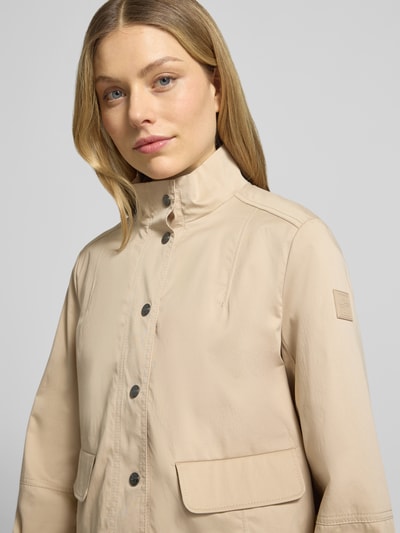 Frauenmodel trägt Regular Fit Jacke mit Stehkragen von Marc Cain in Beige mit Pattentaschen und Logo-Applikation, Nahaufnahme Oberkörper. Elastischer Baumwoll-Mix, elastische Ärmelabschlüsse, Knopfleiste, glatte Struktur.