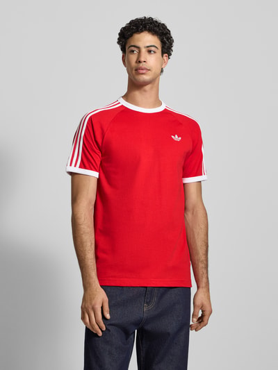 Männermodel trägt T-Shirt mit Label-Stitching von adidas Originals in Rot mit weißen Details, Reine Baumwolle, Regular-Fit, Ganzkörperansicht von vorne.
