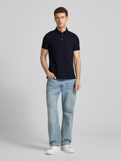 Männermodel trägt Slim Fit Poloshirt mit Stretch-Anteil van Tommy Hilfiger in donkerblauw, korte mouwen, logo, katoen-elastaan-mix, Ganzkörperansicht von vorne.