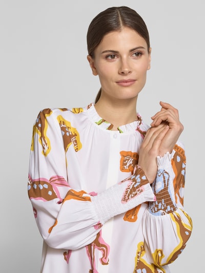 Frauenmodel trägt Bluse mit Rüschen und Smok-Details von HERZENS, Weiß mit buntem Muster, gerader Schnitt, Nahaufnahme Oberkörper. Rundhalsausschnitt mit Rüschen, verdeckter Verschluss, Smok-Details an den Ärmeln, Rückenlänge bei Größe 36: 28 cm, Ärmellänge bei Größe 36: 64 cm.