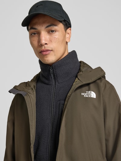 Męski model nosi kurtkę Regular Fit Jacke mit Logo-Print Modell 'Quest' od The North Face, kolor oliwkowy, wpuszczane kieszenie, kaptur ze stójką, zamek błyskawiczny, Szerokość w rozmiarze M: 60 cm, Długość tyłu: 72 cm, Szerokość ramion: 46 cm, Długość rękawów: 69 cm, zbliżenie na górną część ciała.