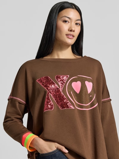 Frauenmodel trägt Oversized Sweatshirt aus reiner Baumwolle Modell 'XO Heartface' von miss goodlife in Braun mit buntem XO-Statement-Print, Ziernähten, elastischen Abschlüssen, Nahaufnahme Oberkörper.