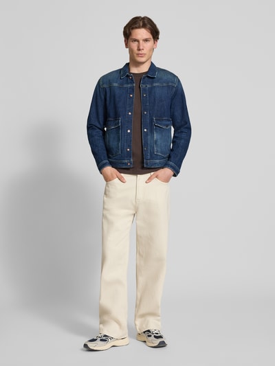 Męski model nosi Jeansjacke mit Pattentaschen und Kentkragen od Replay, mieszanka bawełny z dodatkiem streczu, krój straight fit, ciemnoniebieski denim, metaliczne zatrzaski, detal z logo, kieszenie z patką, zapięcie na zatrzask, długość tyłu w rozmiarze M 68 cm, szerokość ramion w rozmiarze M 38 cm, szerokość klatki piersiowej w rozmiarze M 51 cm, długość rękawów w rozmiarze M 66 cm, jasne spodnie, obuwie sportowe, neutralne tło, pełny widok od przodu.