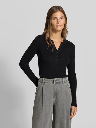 Frauenmodel trägt Slim Fit Longsleeve in Strick-Optik von MYON, schwarz, Henley-Ausschnitt, kombiniert mit grauer Hose, Ganzkörperansicht von vorne.