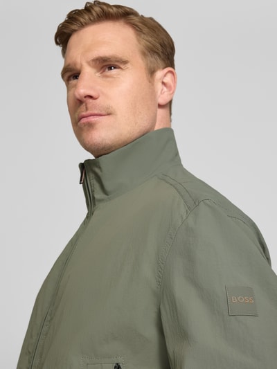 Mannenmodel draagt Relaxed Fit blouson uit waterafstotend materiaal model 'OPENS' van BOSS Orange in effen kleuren, labelpatch, steekzakken, opstaande kraag, tweewegritssluiting, lange mouwen met geribde mouwboorden, rompzoom geribd, zij-ingang steekzakken, detailopname bovenlichaam schuin van voren.