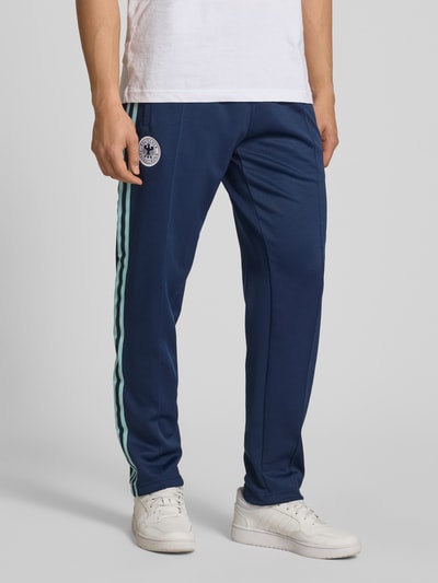 Männermodel trägt Regular Fit Sweatpants mit Vereins Logo Applikation Modell 'BLNACO' von adidas Originals, Optik: Unifarben, Kontraststreifen, Logo-Stitching, fixierte Bügelfalten, elastischer Bundabschluss mit Tunnelzug, Eingrifftaschen, Ganzkörperansicht von vorne.