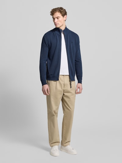 Męski model nosi Cardigan mit Stehkragen und Reißverschluss od Lerros z bawełny, krój regular fit, dwa kolory, wyhaftowane logo, zamek błyskawiczny, długie rękawy, długość tyłu 70 cm, beżowe spodnie, białe buty sportowe, całe ciało widok z przodu.