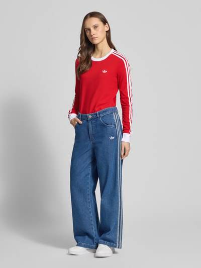 Frauenmodel trägt Longsleeve mit Rundhalsausschnitt und Logo-Stitching von adidas Originals in Rot mit weißen Details, körpernaher Schnitt, Baumwoll-Mix mit Stretch-Anteil, Ganzkörperansicht von vorne.