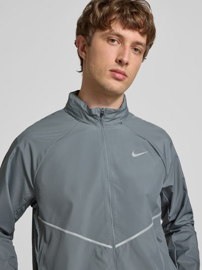 Männermodel trägt Regular Fit Jacke mit Stehkragen Modell 'Miler' von Nike Training in Grau mit Logo-Print, Reißverschluss, Nahaufnahme Oberkörper.