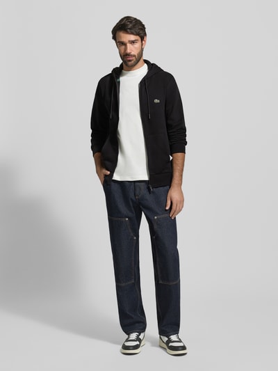 Männermodel trägt Sweatjacke mit Reißverschluss von LACOSTE, Baumwoll-Mix, Regular-Fit, Kapuze, Tunnelzug, Label-Detail, Eingrifftaschen, gerippte Abschlüsse, Rückenlänge 67 cm, Ärmellänge 65 cm, schwarze und blaue Farbtöne, glatte und strukturierte Oberflächen, Ganzkörperansicht von vorne.