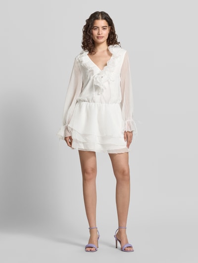 Vrouwenmodel draagt Minikleid met Rüschen und V-Ausschnitt van Gina Tricot in wit, getailleerd model, geplooide details, lange mouwen met vetersluiting, ruglengte maat XS 85 cm, borstwijdte 49 cm, mouwlengte 64 cm, Ganzkörperansicht van voren.