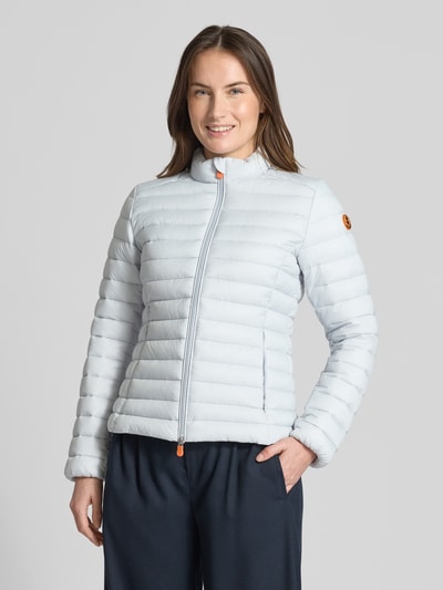 Modelka nosi jasnoszarą Slim Fit Steppjacke gefüttertem Stehkragen Modell 'Carly' od SAVE THE DUCK, struktura w poprzeczne paski, detal pomarańczowy przy zamku, kontekst: damska kurtka pikowana o kroju slim fit, dwukierunkowy zamek błyskawiczny, kieszenie z zamkiem, długość pleców w rozmiarze S: 58 cm, szerokość ramion w rozmiarze S: 39 cm, szerokość w rozmiarze S: 45 cm, długość rękawa w rozmiarze S: 61 cm, widok całej sylwetki z przodu.