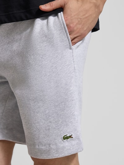 Mężczyznamodel nosi Regular Fit Sweatshorts aus Baumwoll-Mix od Lacoste, jasnoszary, gładka faktura, naszywka z logo, krój regular fit, materiał bawełniany, kieszenie boczne, zbliżenie na górną część nogi.