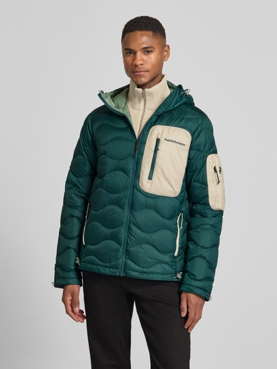 Männermodel trägt Daunenjacke mit Kapuze von PEAK PERFORMANCE in Grün mit beigen Reißverschlusstaschen, gerader Schnitt, Ganzkörperansicht von vorne.