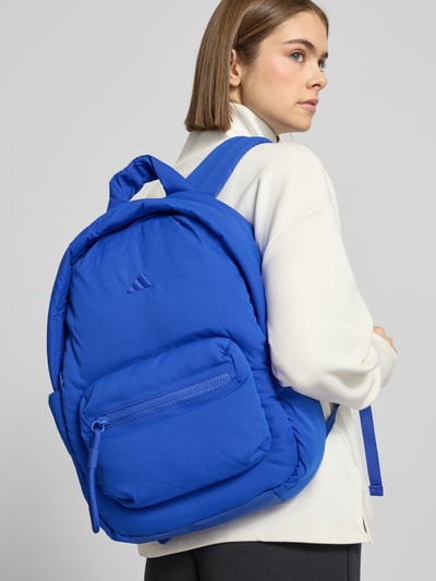 Frauenmodel trägt Rucksack mit Tragehenkel von adidas Sportswear in kräftigem Blau mit Hauptfach, Reißverschluss, Label-Detail, Nebenfach, Ganzkörperansicht Rückansicht