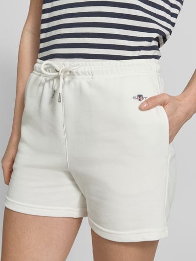 Modelka kobieca ma na sobie Sweatshorts mit Eingrifftaschen und Label-Stitching od Gant, biały kolor, bawełniany miks, krój regular fit, elastyczny pas, kieszenie, haftowane logo, Szerokość w rozmiarze S: 36 cm, długość wewnętrzna nogawki S: 10 cm, zdjęcie z przodu, zbliżenie na górną część ciała.
