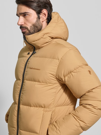 Männermodel draagt gewatteerd jack met capuchon, labelstitching, tunnelkoord, ritssluiting, steekzakken, van Peak Performance, regular fit, kleur beige, context: Heren gewatteerd jack Steppjacke mit Kapuze und Label-Stitching van PEAK PERFORMANCE, regular fit, labelstitching, capuchon met tunnelkoord, ritssluiting, steekzakken, ruglengte maat M 72 cm, schouderbreedte 49 cm, borstwijdte 52 cm, mouwlengte 70 cm. Detailaufnahme.