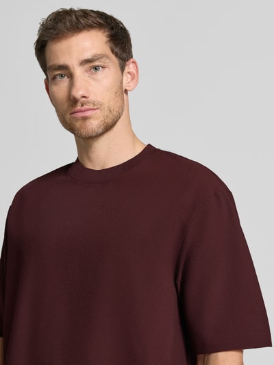 Männermodel trägt T-Shirt mit Strukturmuster und Rundhalsausschnitt von Emporio Armani in Dunkelrot, Material-Mix, Nahaufnahme Oberkörper.
