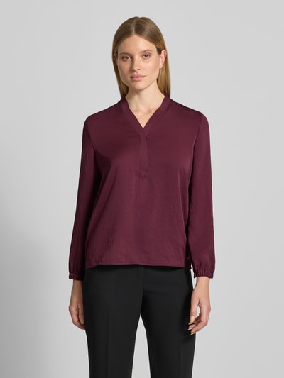 Kobiece model nosi Regular Fit Bluse mit V-Ausschnitt od Marc Cain, krój regular fit, dekolt w serek, detal z logo, długość tyłu w rozmiarze 36: 65 cm, długość rękawów w rozmiarze 36: 59 cm, kolor bordowy, gładka faktura, widok całej sylwetki z przodu.