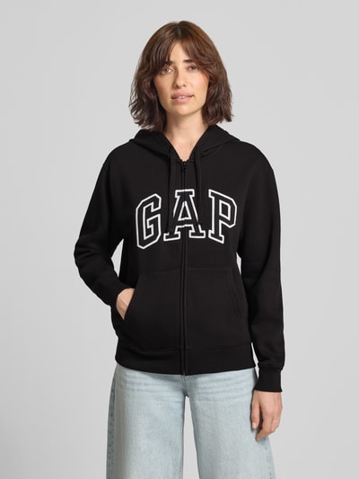Vrouwenmodel draagt zwarte sweatjack met capuchon en kangoeroezak Model 'HERITAGE' van GAP, katoenmix, relaxed fit, logoprint, ritssluiting. Ganzlichaanzicht van voren.