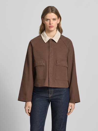 Frauenmodel trägt Regular Fit Jeansjacke aus reiner Baumwolle Modell 'BETT' von Pieces, brązowy jednokolorowy denim, wykładany kołnierz z obszyciem ze sztruksu, krój regular fit, długi rękaw, raglanowe rękawy, kieszenie z patką, listwa z zatrzaskami, wykończenie: denim, materiał: bawełna, ganzkörperansicht von vorne.