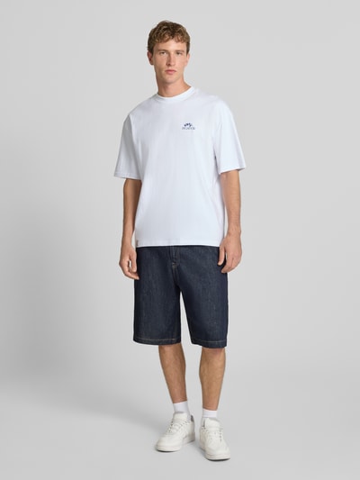 Männermodel trägt Oversized T-Shirt mit Backprint von PEGADOR aus weißer Baumwolle, kombiniert mit Jeansshorts, Ganzkörperansicht von vorne.