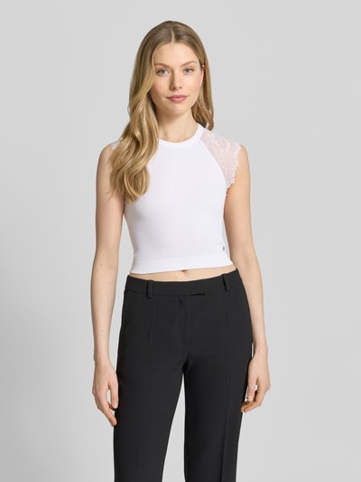 Modelka kobieca nosi Crop Top mit Spitzenbesatz Modell 'MILLIE' od Guess, biały materiał z koronką, krój slim fit, długość 37 cm w rozmiarze S, ujęcie całej sylwetki z przodu.