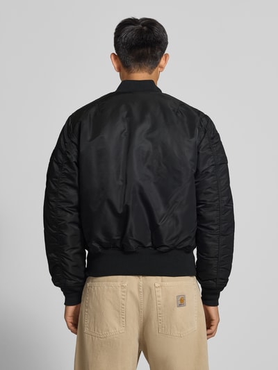 Männermodel trägt schwarze Bomberjacke mit Pattentaschen und Label-Applikation von Alpha Industries, Ganzkörperansicht Rückansicht. Regular Fit, Stehkragen, Reißverschluss, Rücklänge 69 cm, Schulterbreite 44 cm, Brustweite 54 cm, Ärmellänge 64 cm.