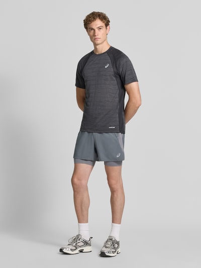 Męski model nosi Regular Fit Shorts mit integrierten Tights Model 'Road' von ASICS TIGER, kolor szary, detal z logo, krój regular fit, elastyczny pas, Szerokość w rozmiarze M: 38 cm, Długość wewnętrzna nogawki: 12 cm; zdjęcie całej sylwetki z przodu.