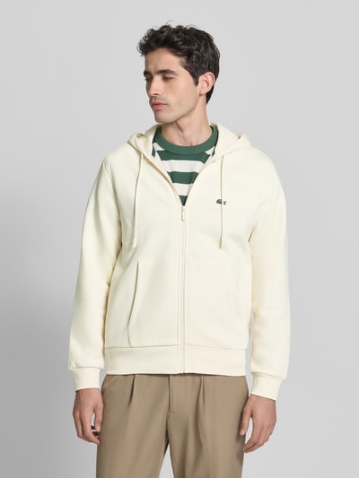 Männermodel trägt Regular Fit Sweatjacke aus Baumwoll-Mix von Lacoste in Optik: Unifarben mit Reißverschluss, Logo-Stitching, Kapuze mit Tunnelzug, Langarm, geripptem Ärmel- und Rumpfabschluss, geteilter Kängurutasche, Materialverarbeitung: Sweat, Farbe creme, Textur weich, Ganzkörperansicht von vorne.