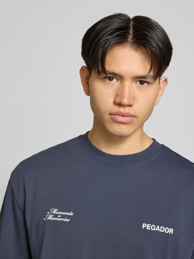 Mężczyzna nosi Oversized T-Shirt mit Label-Detail Modell 'Rochefort' od Pegador, czysty bawełniany materiał, ciemnogranatowy kolor, nadruk z przodu, okrągły dekolt, motyw na plecach, kontekst: męska koszulka Pegador, szeroki krój, longline, długość pleców w rozmiarze M: 72 cm, zbliżenie na górną część ciała
