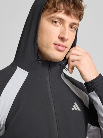 Männermodel trägt Sweatjacke mit Kapuze von adidas Training in Schwarz-Grau mit Material-Mix, Stretch-Anteil, Label-Print, Reißverschluss, Nahaufnahme Oberkörper.